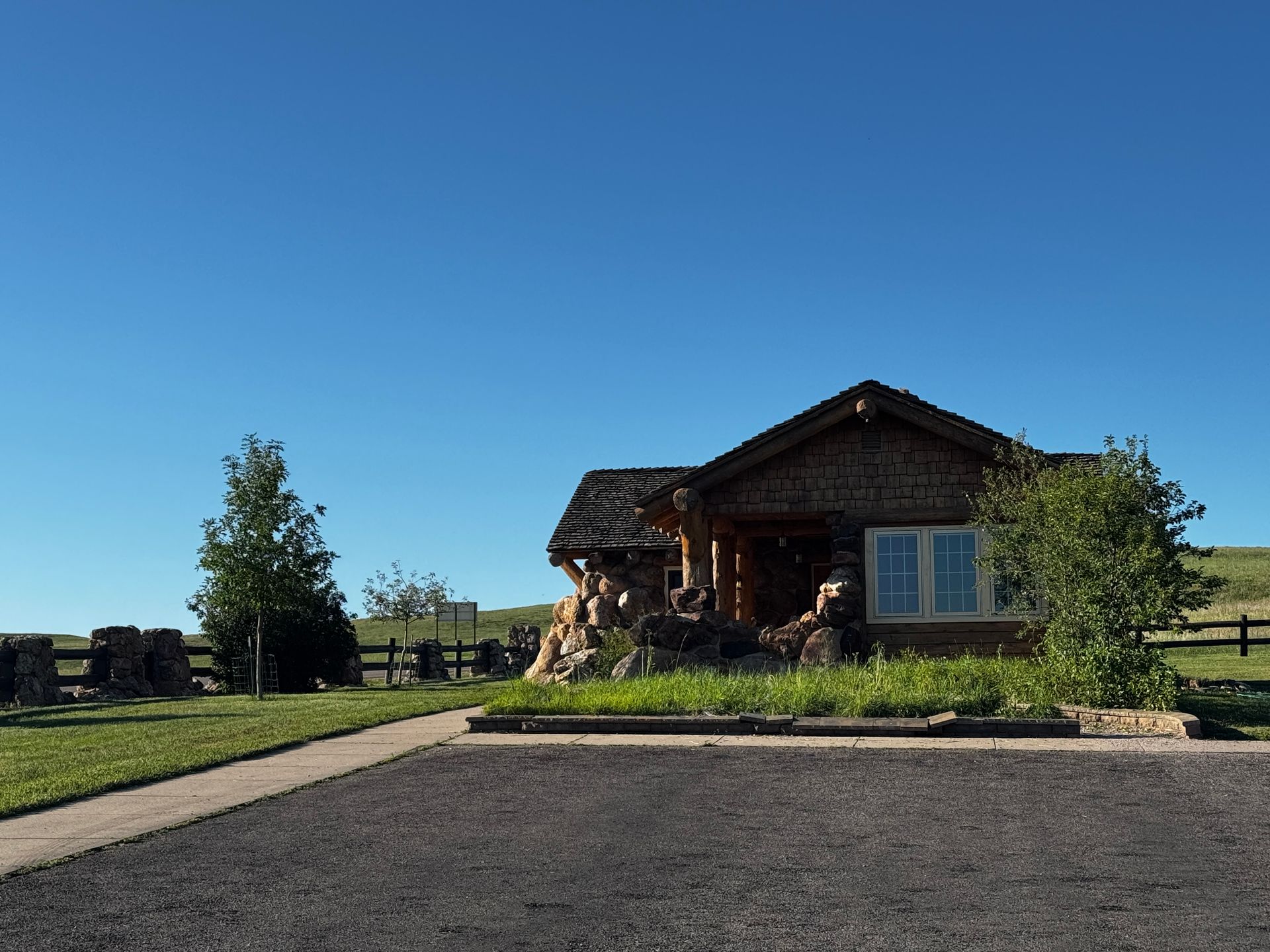 Log cabin with stone facade under a clear, blue sky; green lawn, asphalt parking area.