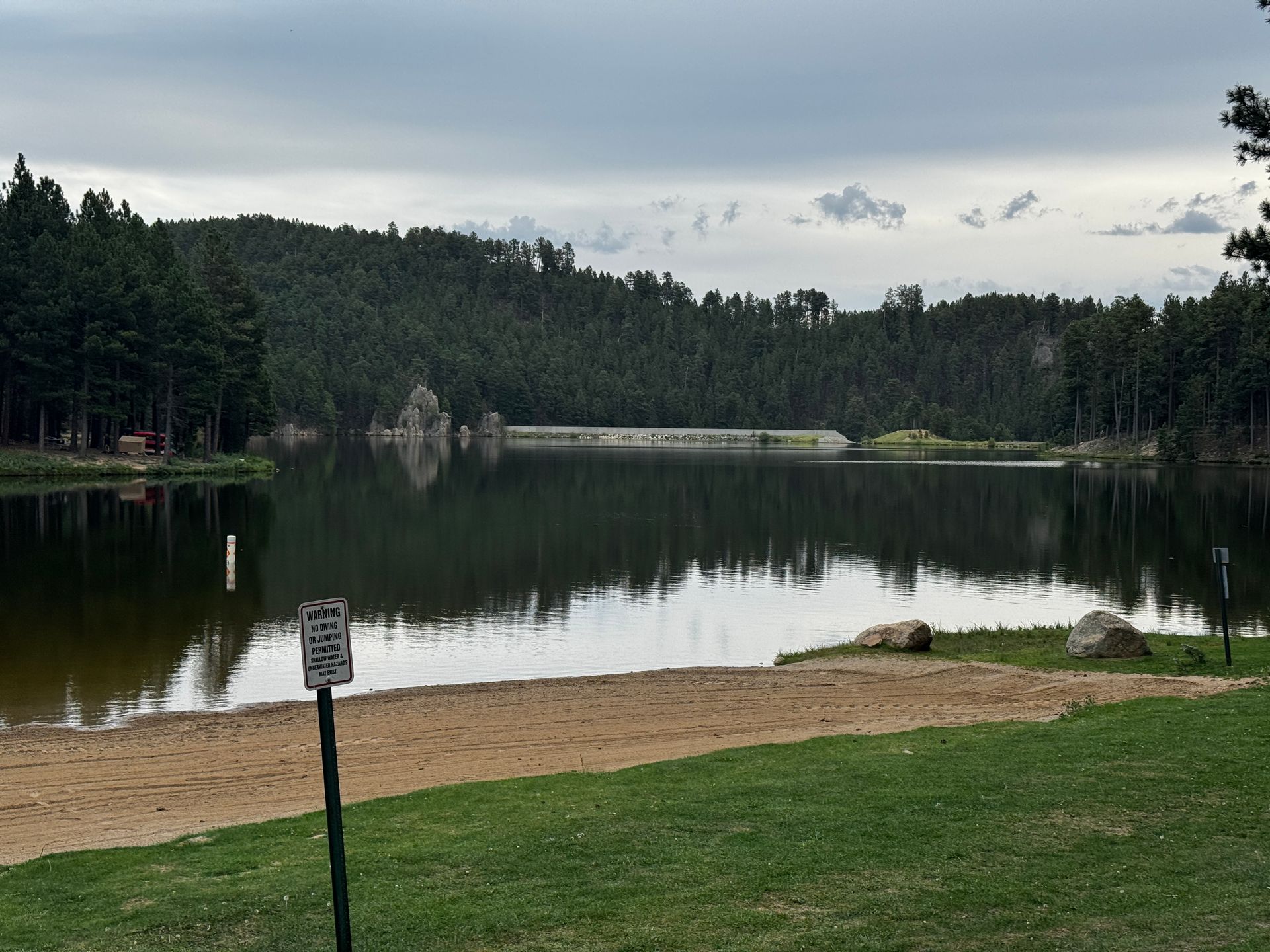 Lake with trees, dam, and overcast sky; sandy shore in foreground.