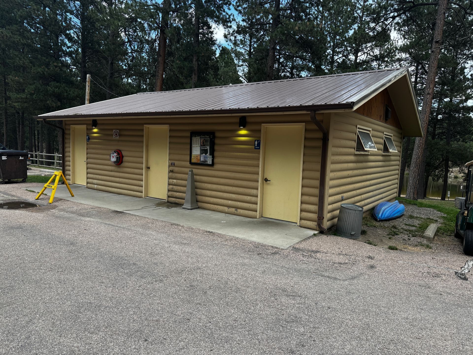 A log cabin-style public restroom building with yellow doors, outdoor lighting, and gravel parking.