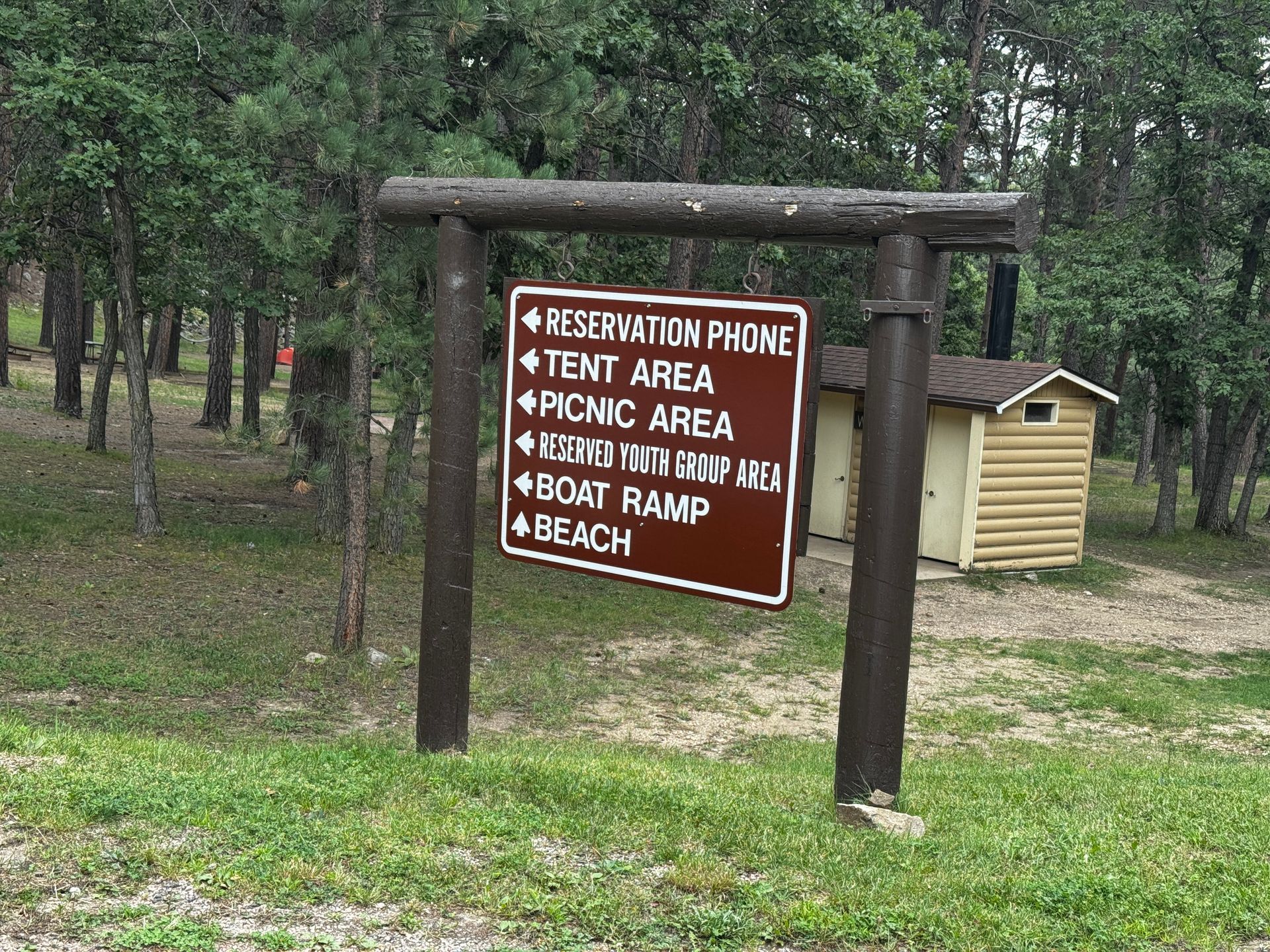 Signpost indicating amenities: reservation phone, tent area, picnic area, beach, boat ramp in a wooded area.