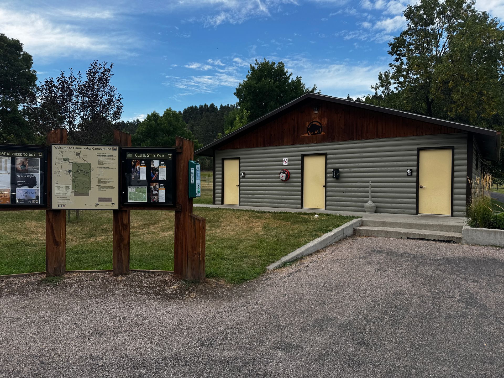 Log cabin public restroom with information kiosk, gravel parking, grass, and trees under a blue sky.
