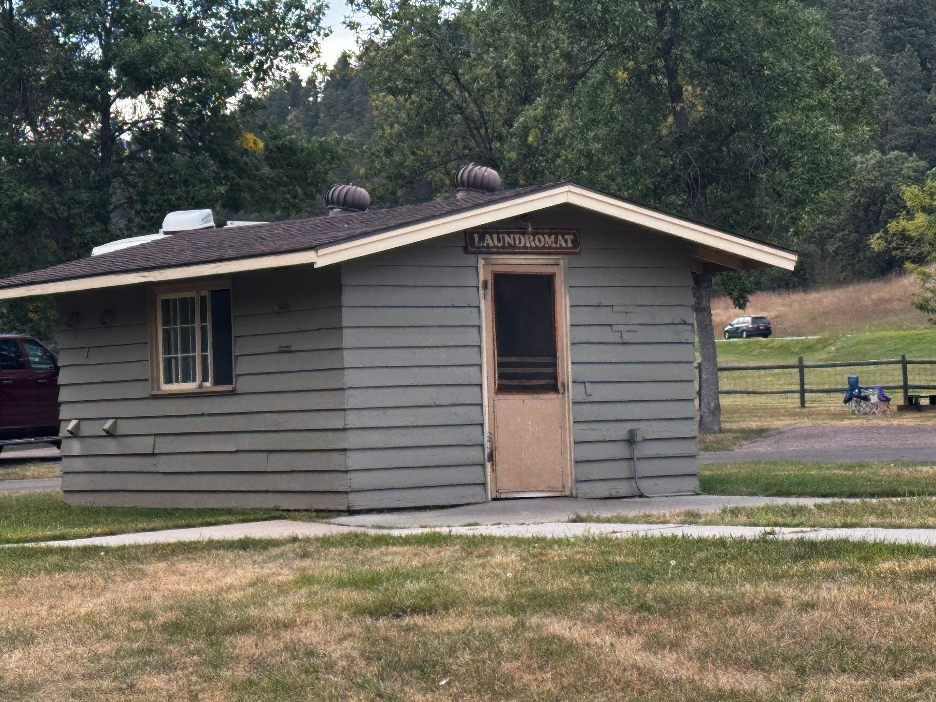 Small, weathered gray building with a tan door and sign above it, in a grassy outdoor area.