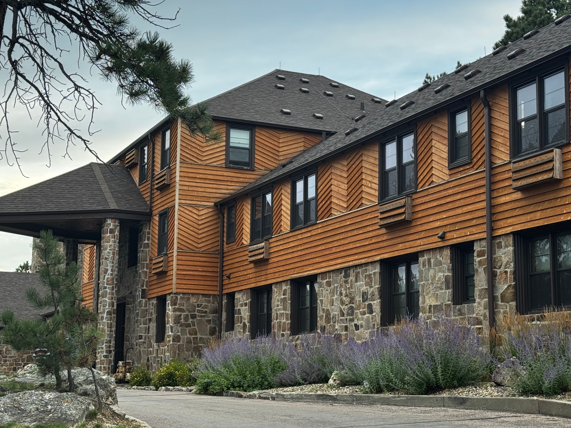 A multi-story lodge with wood and stone facade, brown roof, and lavender bushes along the driveway.