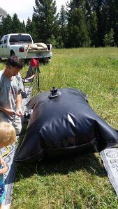 Man inflating a large black water bladder in a grassy field near a white truck.