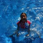 Girl in pink swimsuit smiles in bright blue pool water.
