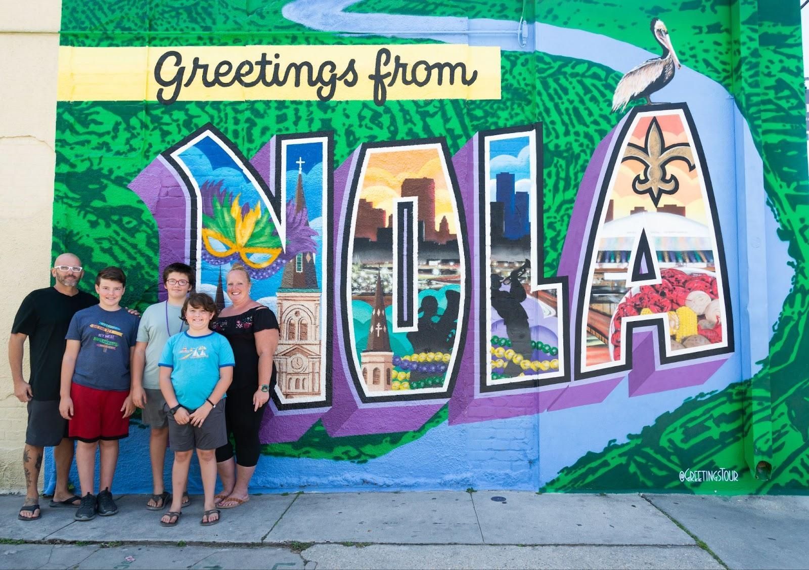 A family standing in front of a mural that says greetings from nola