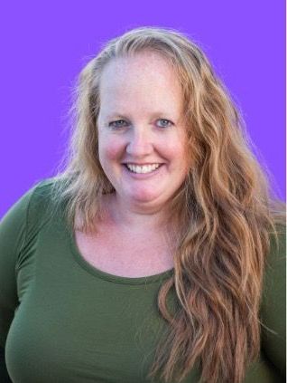 Woman with long wavy blonde hair smiles, wearing an olive green long-sleeved shirt against a purple background.