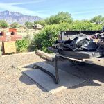 A vehicle's open tailgate with debris and a curved metal bar on a cement board in a sunny outdoor setting.