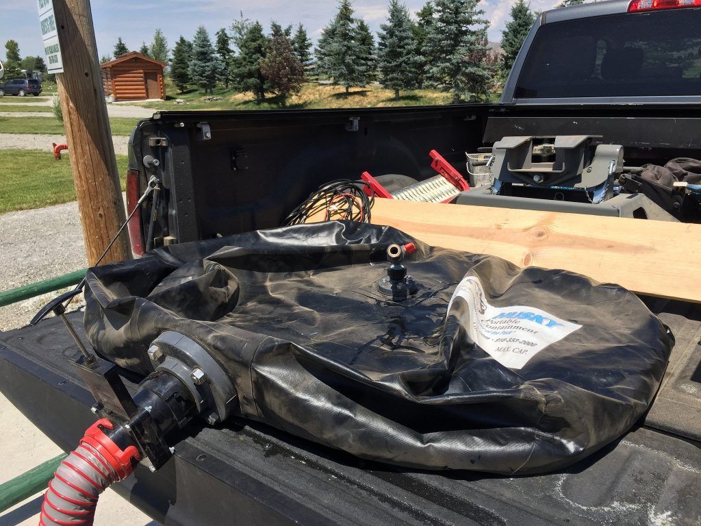 Black water tank in the bed of a truck, with a hose attached. Outdoors, on a sunny day.