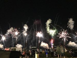 Fireworks exploding in night sky over a crowd of people and cars, creating white and colorful bursts.