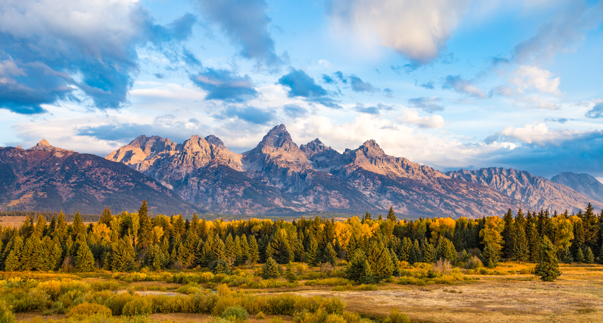 Mountains rise above a forest and field under a partly cloudy sky.