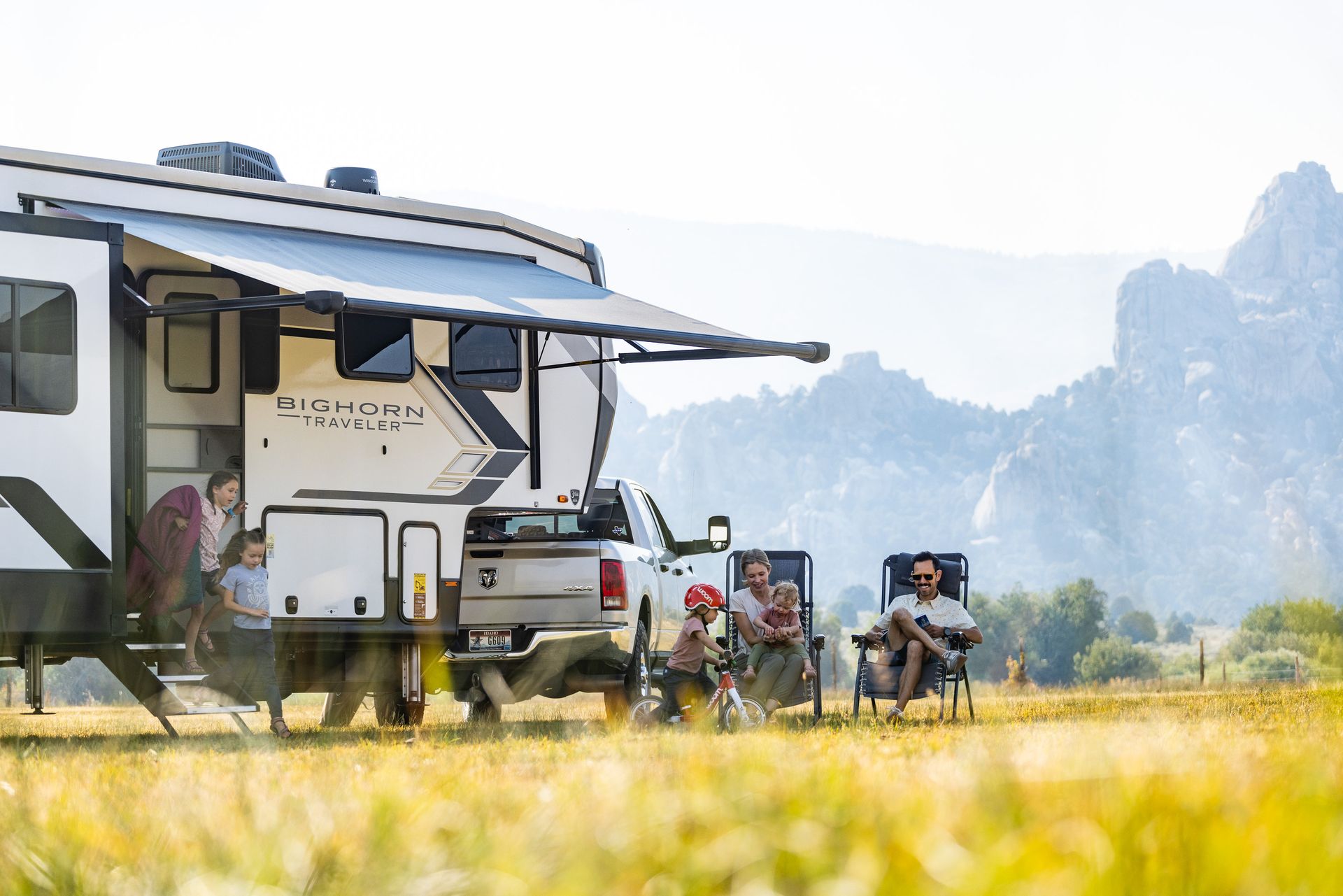RV campsite with people relaxing in foldable chairs beside a white camper, mountains in the background
