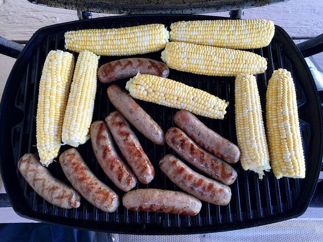 Corn on the cob and sausages are cooking on a grill.