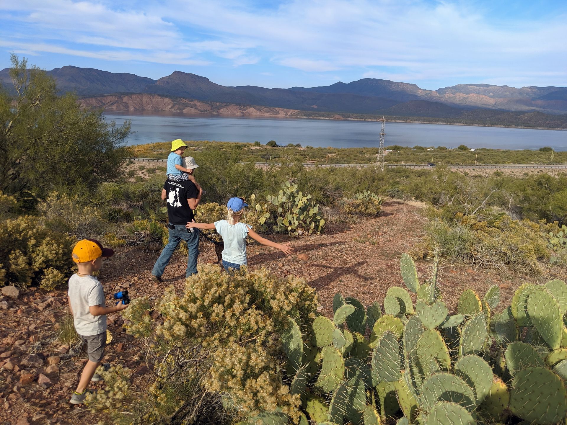 Family hiking near a lake, sunny day. One child carried on shoulders. Cacti in foreground, mountains in the distance.