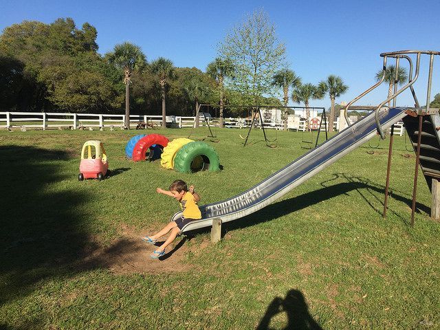 Child slides down metal playground slide in grassy park. Other play equipment visible.