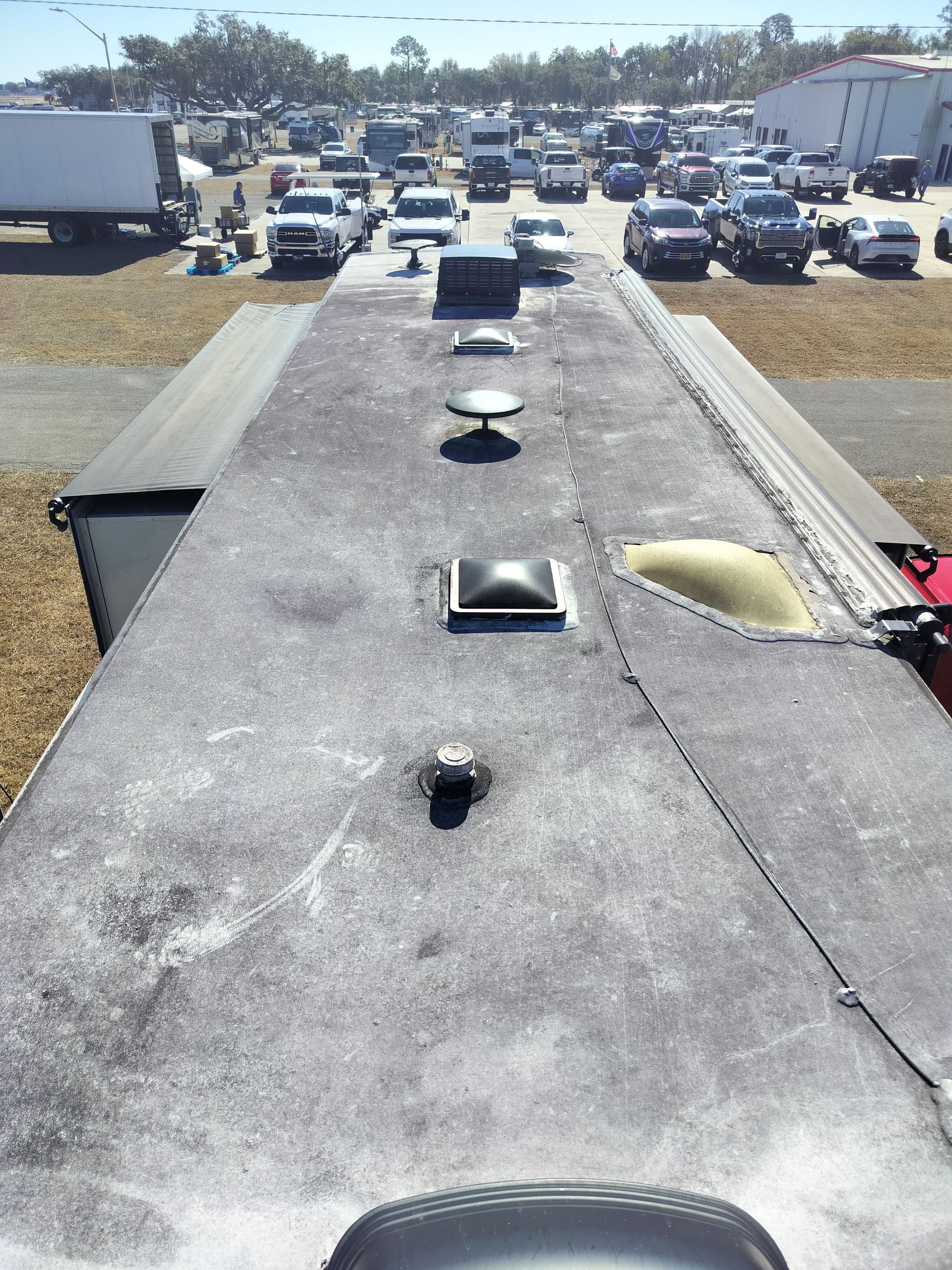 A view looking down the length of a weathered RV roof featuring multiple vents and a skylight in a crowded parking lot.