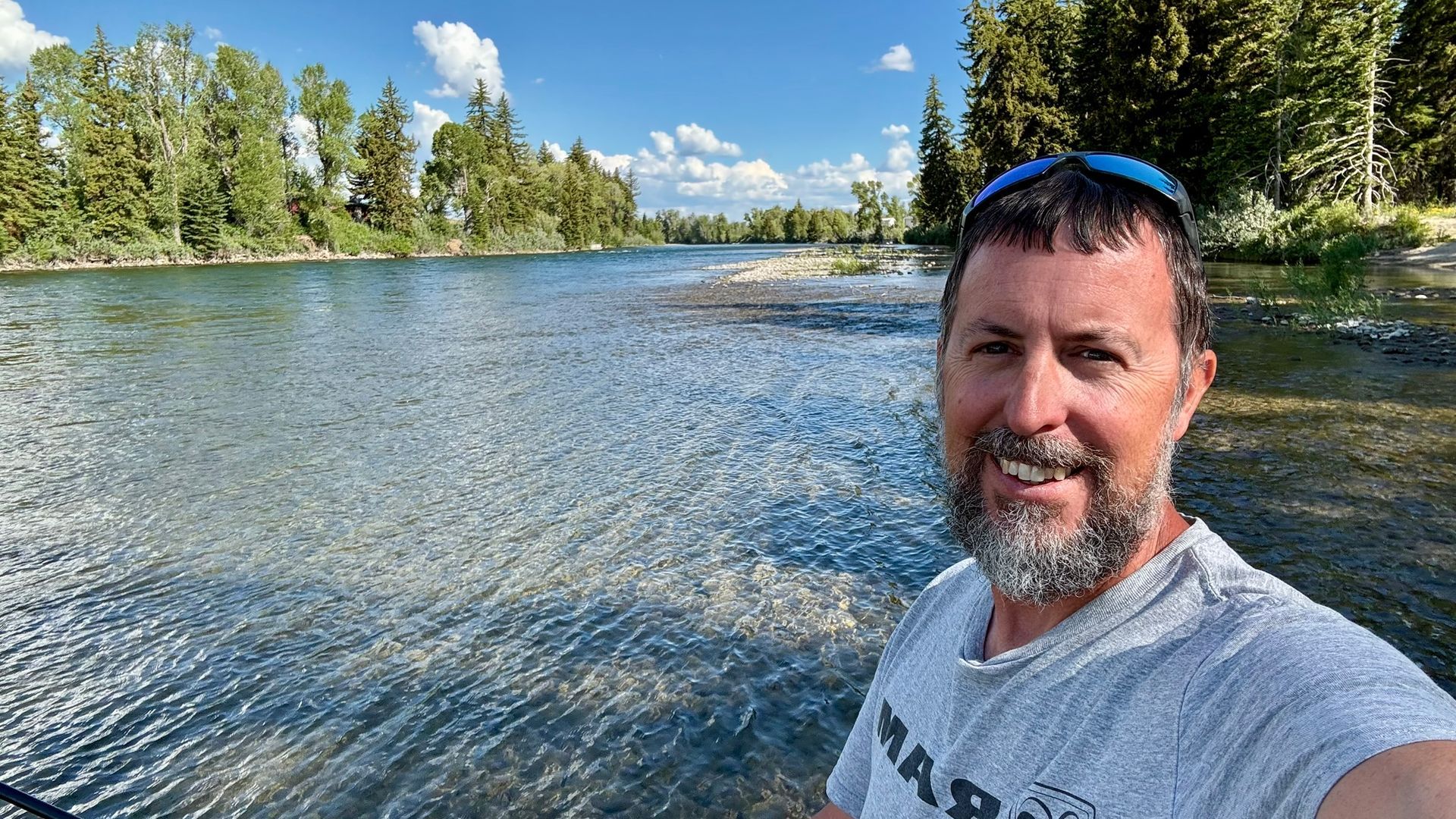 Man smiling by a clear river. Green trees line the bank, blue sky overhead.