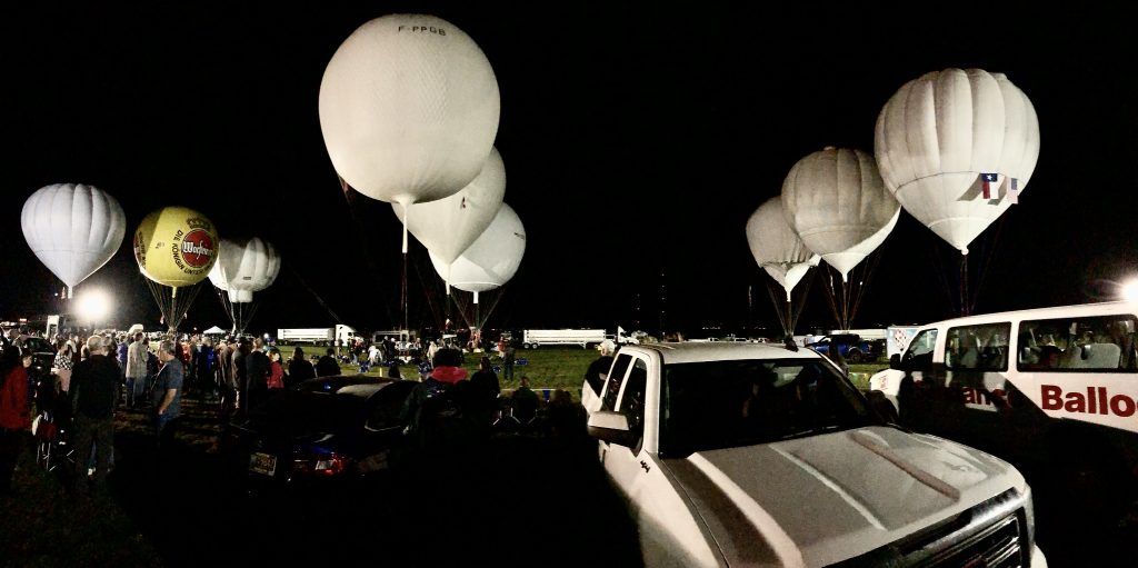 Hot air balloons at night, people in foreground, illuminated by lights.