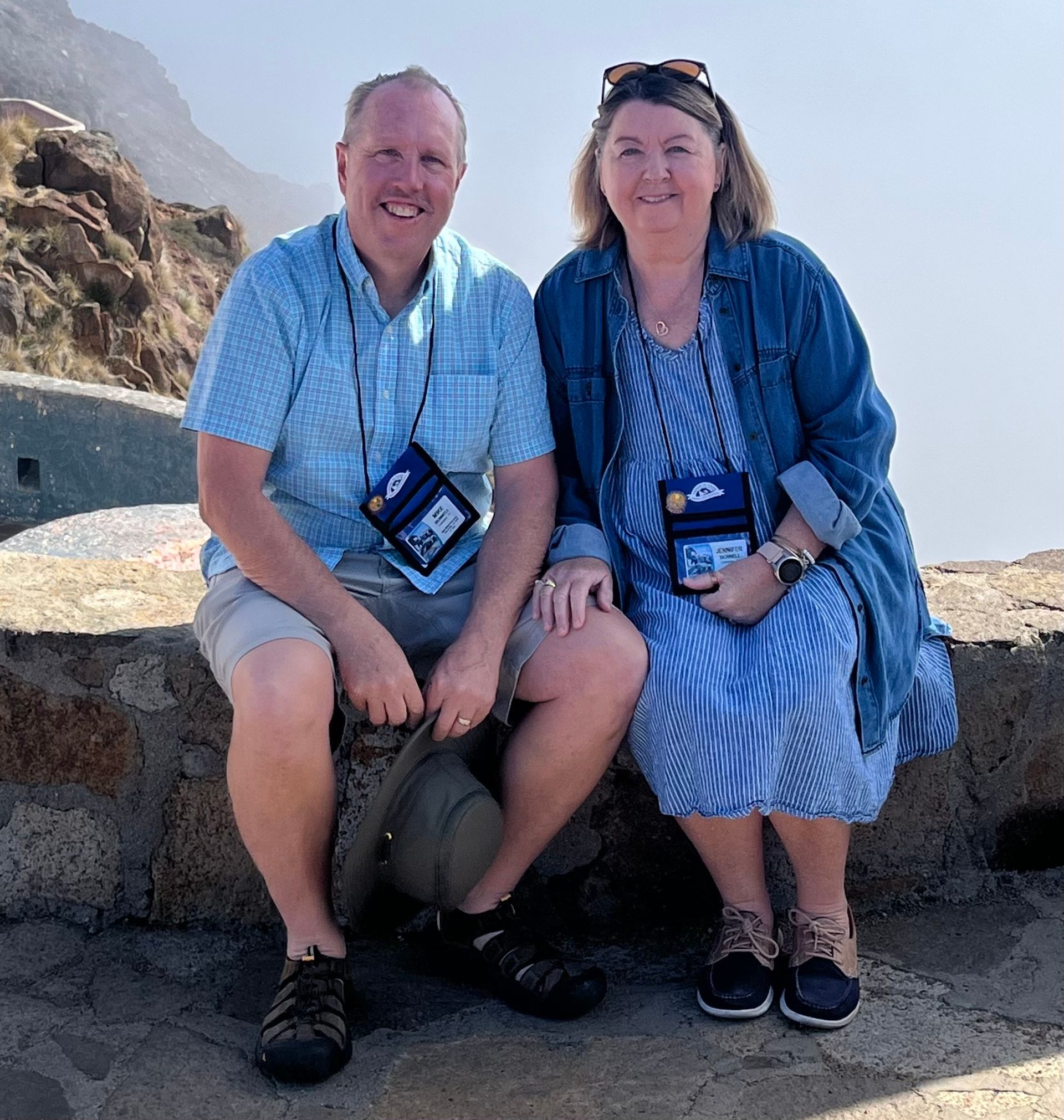 Man and woman seated on a stone wall overlooking a hazy landscape. Both wear lanyards, smiles.