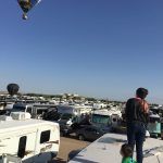 Hot air balloons rise over a crowded RV park under a blue sky. A person and child stand on an RV roof.