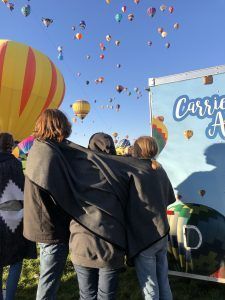 People watch hot air balloons in a blue sky. Balloons of various colors fill the air. A sign says
