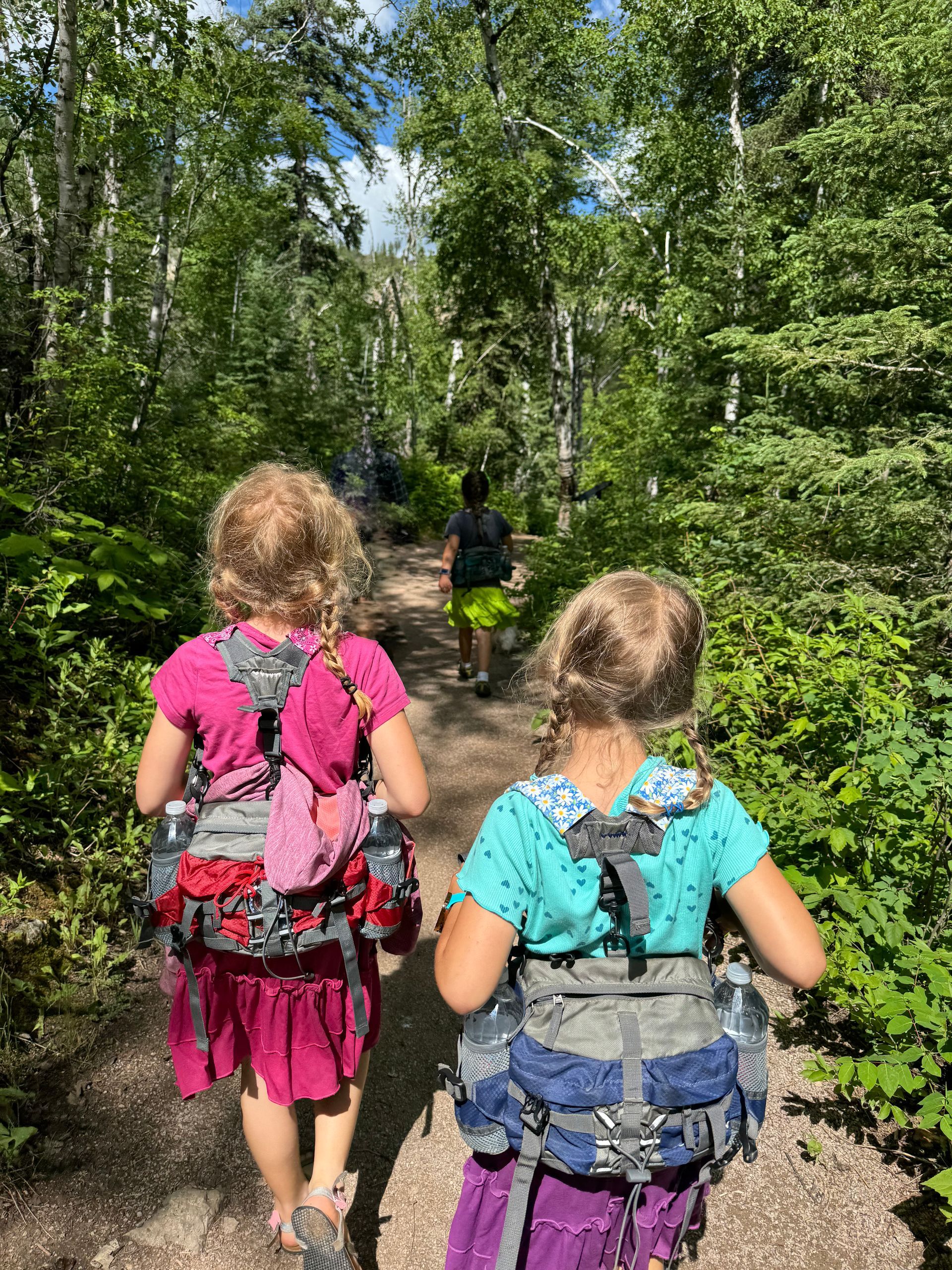 Two girls with backpacks hiking a wooded trail, followed by another person.