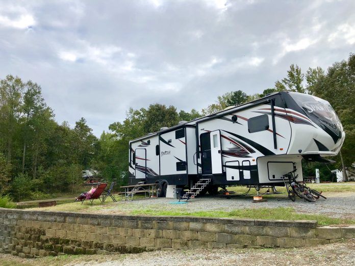RV parked on a grassy patch near trees and a cloudy sky, with picnic table and bikes present.