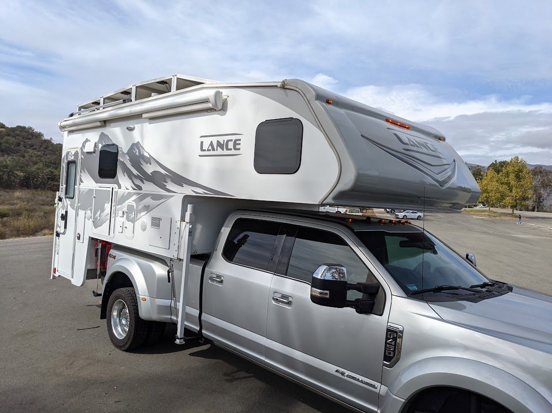 Silver pickup truck with a Lance camper, parked outdoors under a cloudy sky.