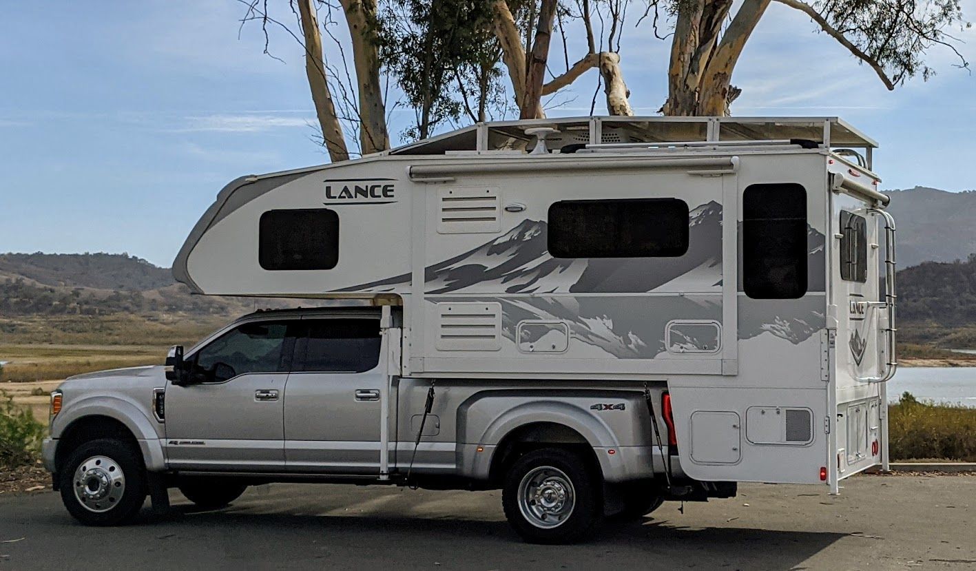 Silver pickup truck with a white Lance camper parked outdoors, near a lake.