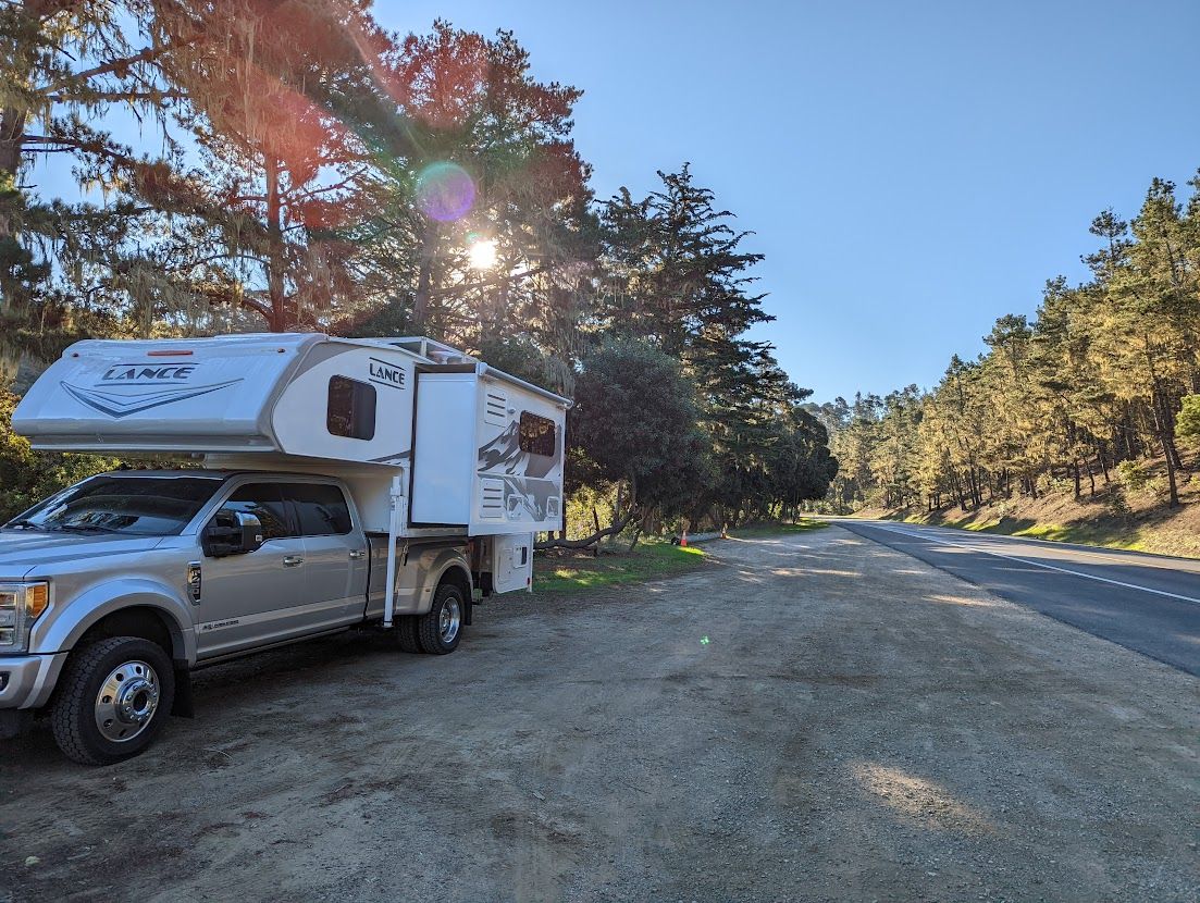 Truck with camper parked on dirt shoulder next to a road, sun shining through trees.
