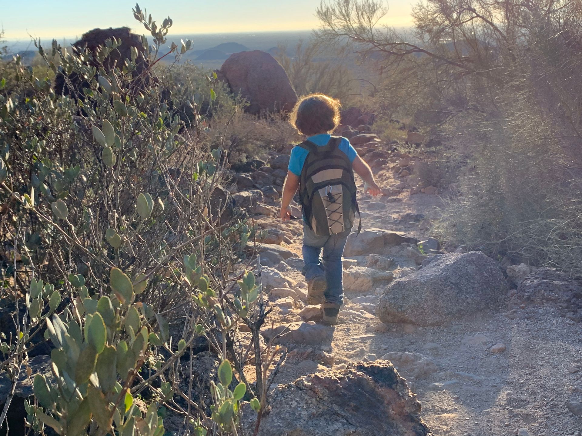 Child hiking on a rocky desert trail, wearing a backpack, with sunlight in the background.