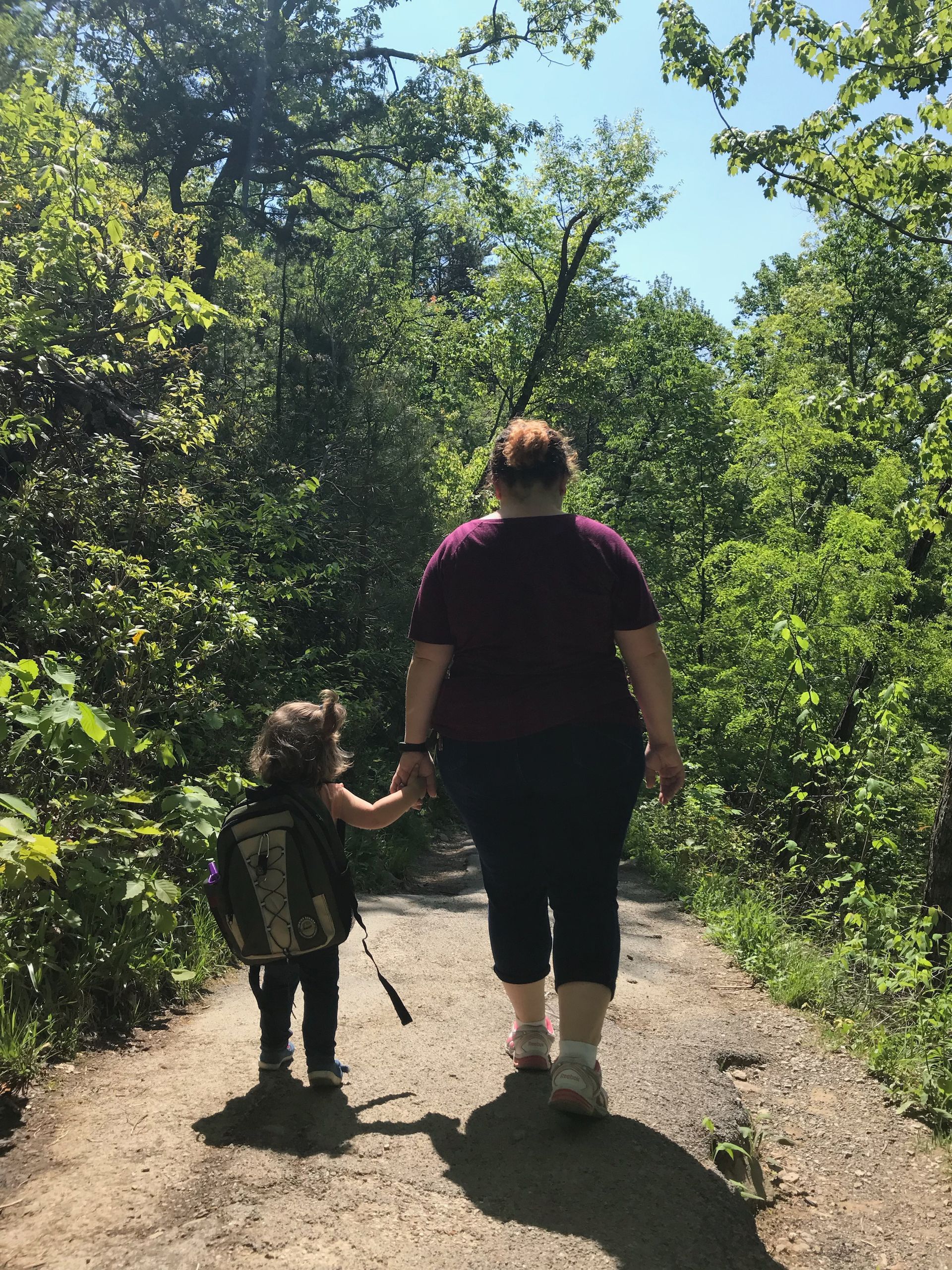 Adult and child hold hands, walking on a path through a forest. Sunlight filters through the trees.