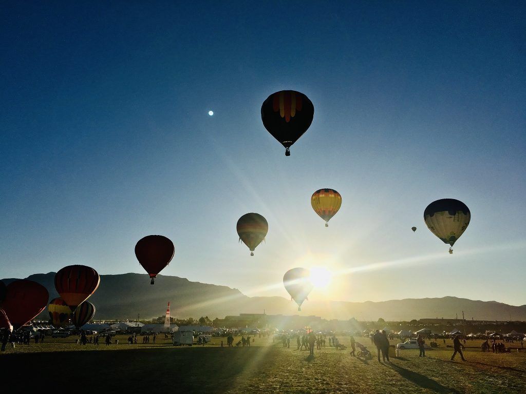 Hot air balloons ascend against a bright sunset over a field with people and mountains.