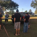 People gathered outdoors, looking at a truck. One man holds a circular object, likely filming a scene.