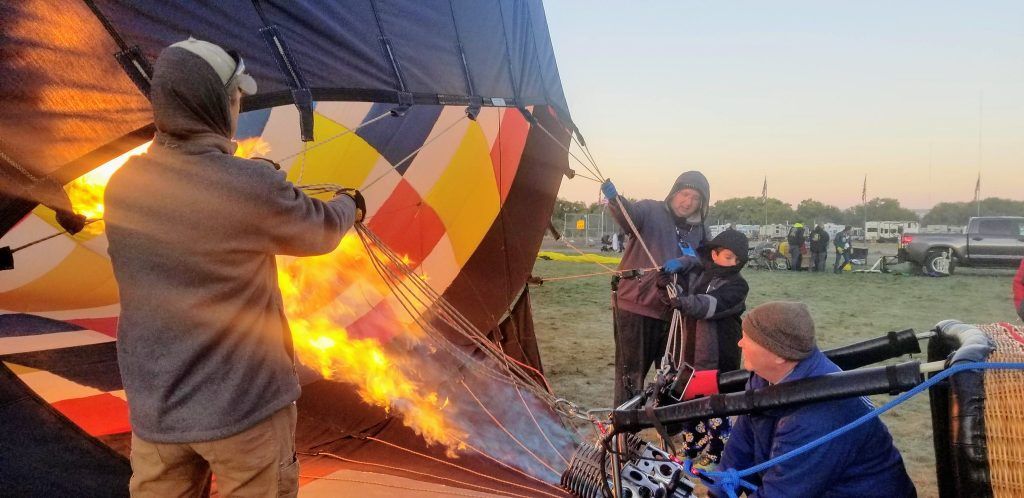 A hot air balloon inflates with fire. People stand nearby, watching the process.