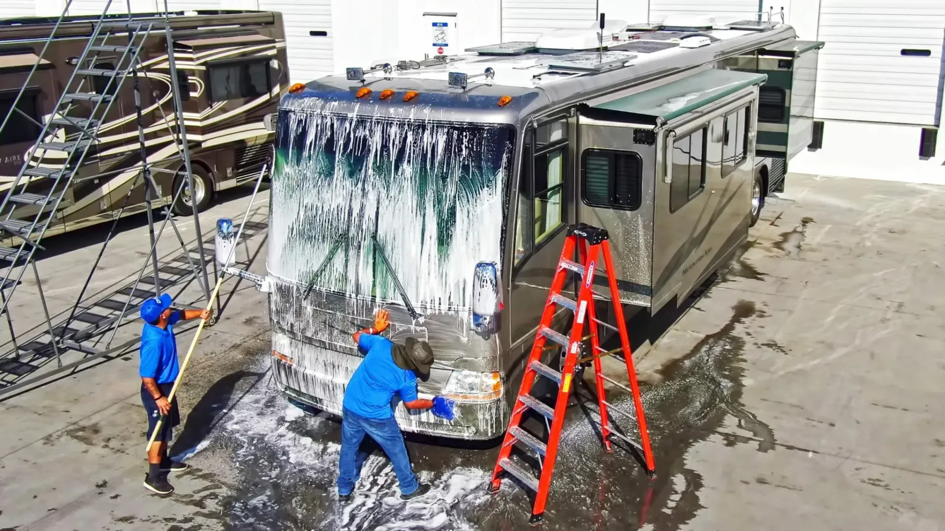 Two people washing a large RV with soap and water outside a building.