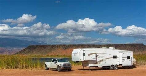 White truck towing a white fifth-wheel trailer on a dirt road by a body of water, under a blue sky with clouds.