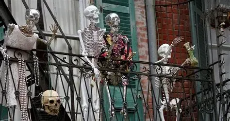 Skeletons in Halloween costumes on a balcony with ornate iron railings, against a brick building.
