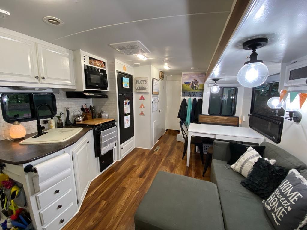 RV interior: kitchen with white cabinets, wooden floors, seating area with gray couch, and dining table.