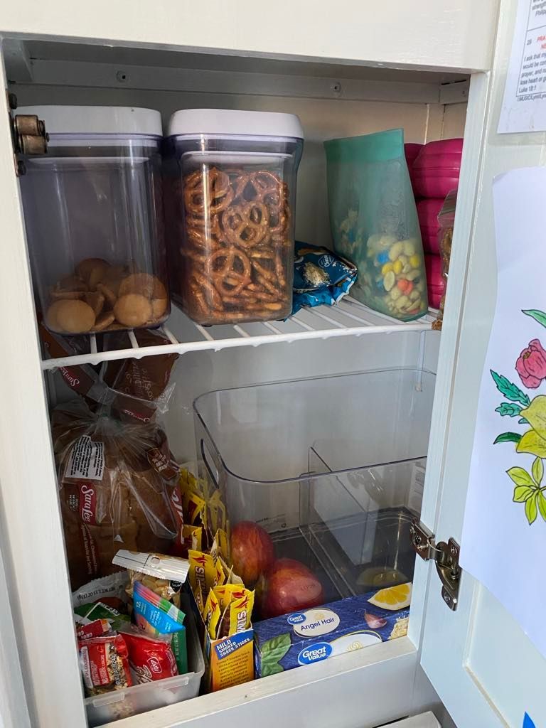A pantry cabinet with food items inside. It holds pretzels, apples, bread, and snacks.