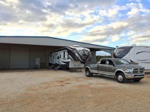 Gray pickup truck towing a large, black and white RV, parked near a building.