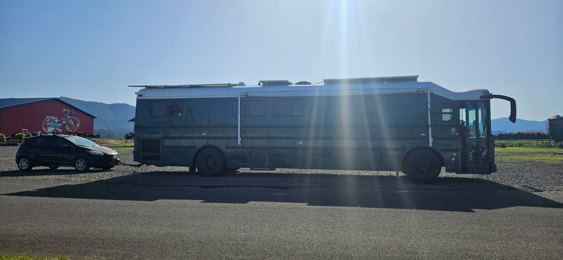 A large gray bus parked on a gravel lot next to a black car and a red barn on a sunny day.