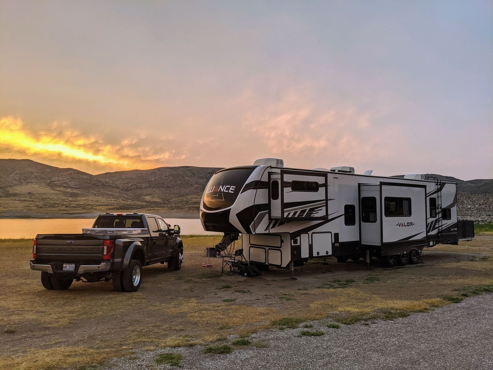 Truck towing a large RV parked near a lake at sunset. The sky has orange and yellow hues.