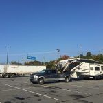 Truck towing a large RV in a parking lot with a semi-truck and blue sky.