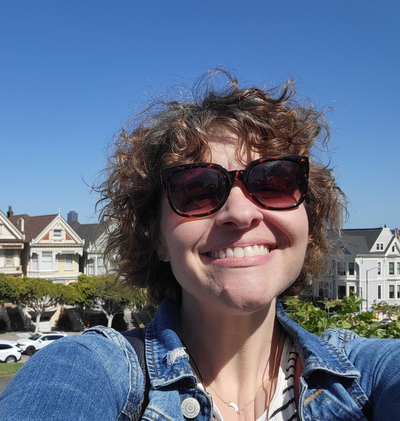 Woman in sunglasses smiles in front of colorful Victorian houses on a sunny day.