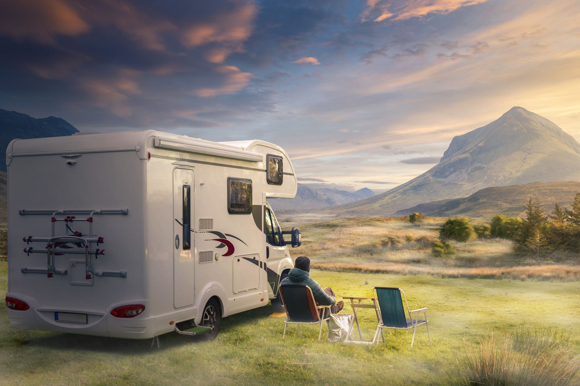 RV parked in a field with a person in a chair enjoying mountain views at sunset.
