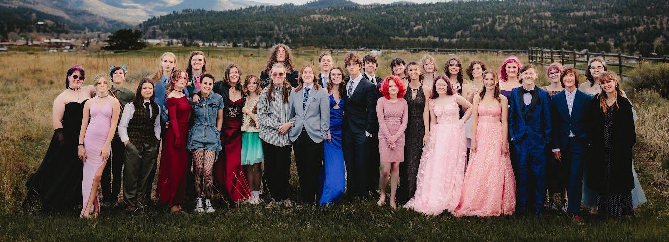 Group of people posing outdoors, many in formal wear, green field and mountain backdrop.