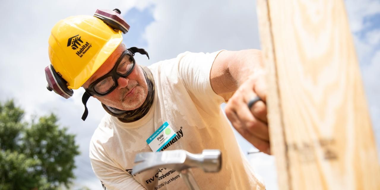 A person wearing safety gear hammers a nail into wood, outdoors.