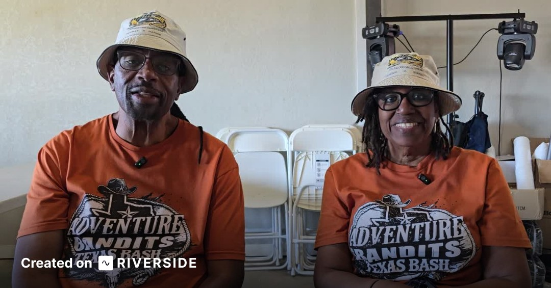 Two people in matching orange shirts and hats sit indoors, smiling at the camera.