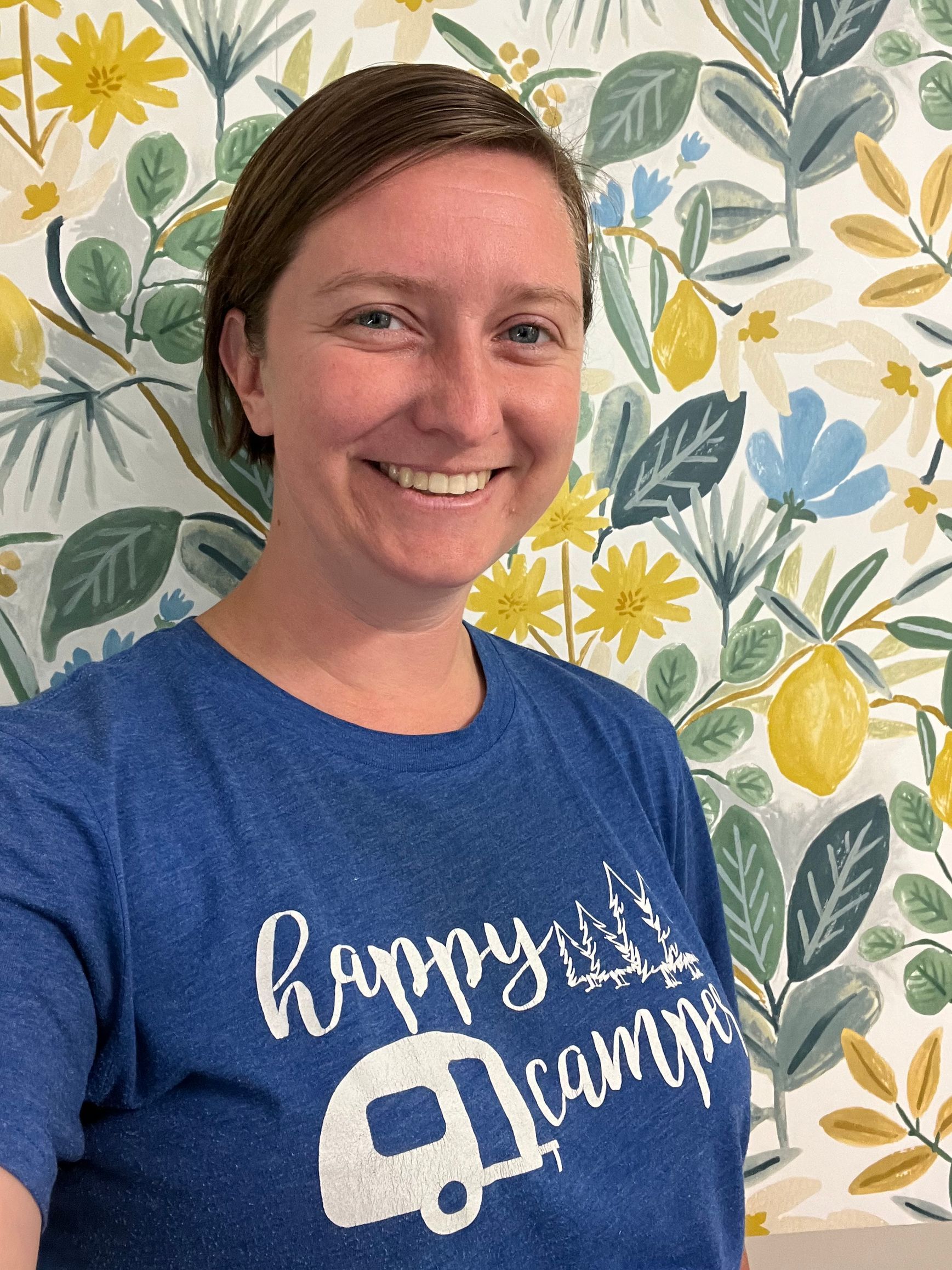 Woman in blue shirt smiling in front of floral wallpaper that has lemons. Shirt says